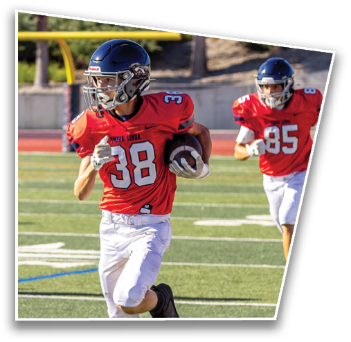 Two people in red football uniforms and helmets are running on a football field with one holding a football. Description generated by AI