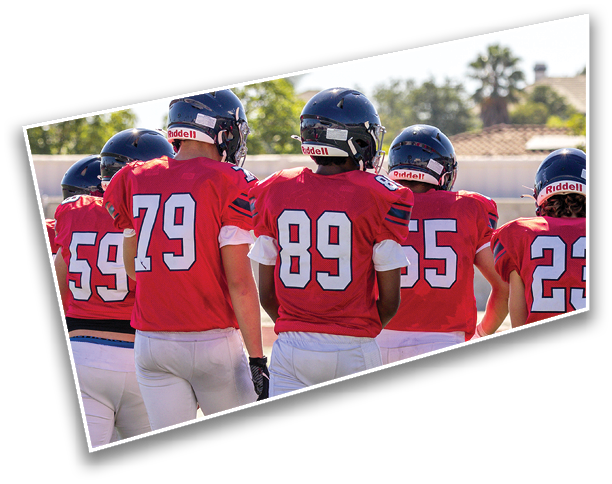 A group of people wearing football helmets and red jerseys are gathered outdoors with trees and a clear sky in the background. Description generated by AI