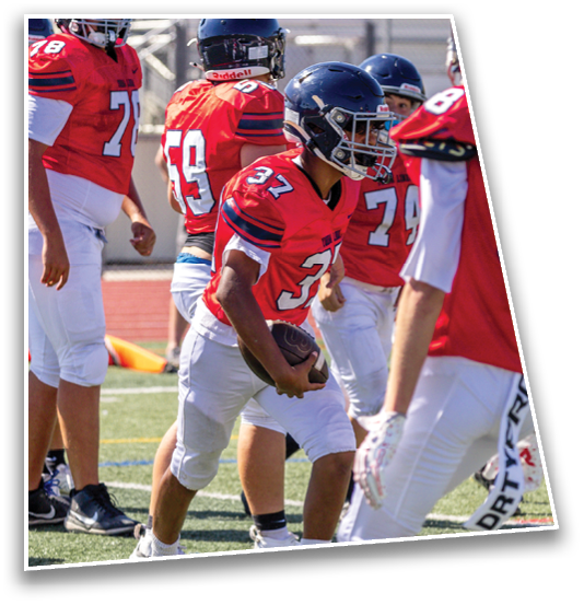 A group of people wearing red football jerseys and white pants are on a field with one person holding a football and preparing to run. Description generated by AI