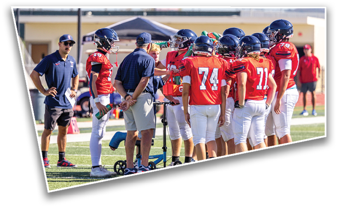 A group of people wearing dark sports uniforms and helmets are standing on a football field near the sidelines, some gesturing and interacting with each other. Description generated by AI