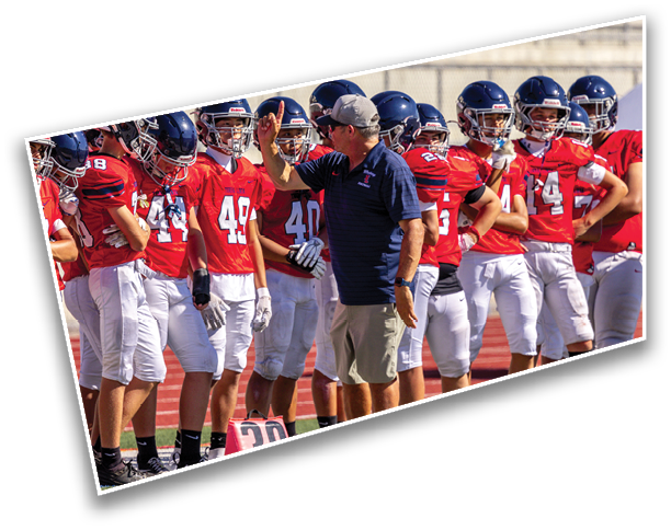 A coach in a cap and navy shirt is giving instructions to a group of football players wearing red jerseys and helmets on a sports field. Description generated by AI
