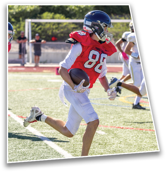 A person wearing a red football jersey with the number 88 is running on a field while holding a football, with other players and a goalpost visible in the background. Description generated by AI