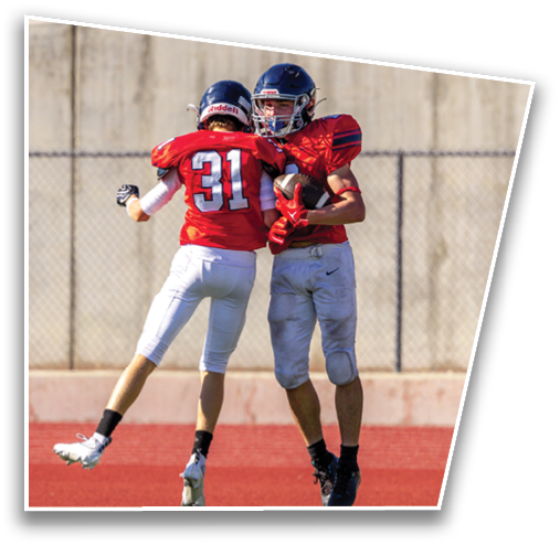Two people wearing red football uniforms and helmets are engaged in a conversation or interaction on a field with a concrete wall and grassy area in the background. Description generated by AI