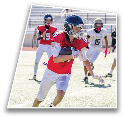 A person in a red football jersey and white pants is running with a football while two other players in white jerseys and one in a red jersey are behind on a sunlit field. Description generated by AI
