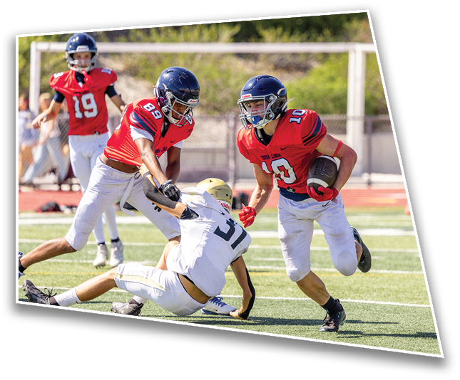 Three adults in football uniforms are playing on a field with one player in white being tackled by two players in red while another player in red watches in the background. Description generated by AI
