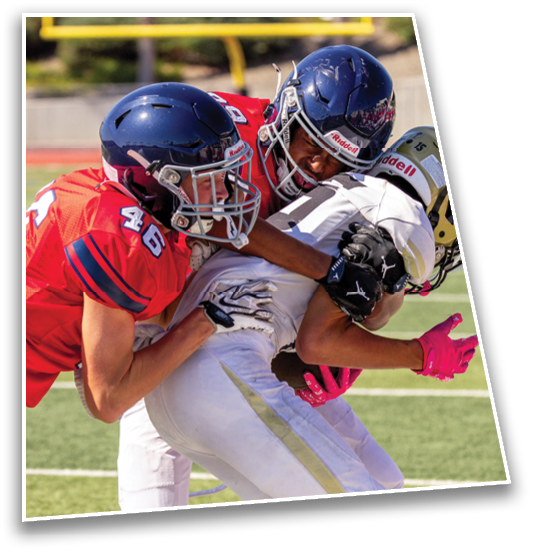 Two people in red football uniforms tackle a person in a white football uniform on a field during a game. Description generated by AI
