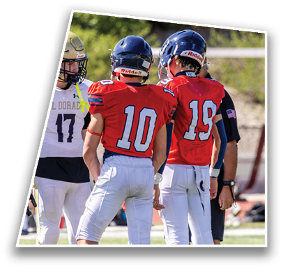 Two people in red football uniforms with helmets stand on a field facing away from the camera while another person in a white uniform and helmet stands nearby. Description generated by AI