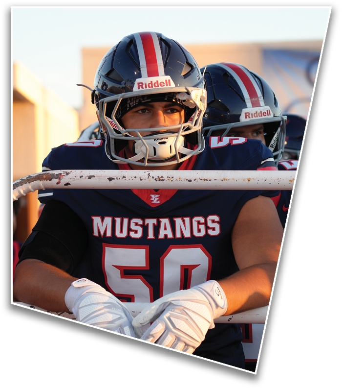 Three people wearing navy blue football uniforms with red and white accents and helmets stand together on a field with a concrete structure in the background. Description generated by AI