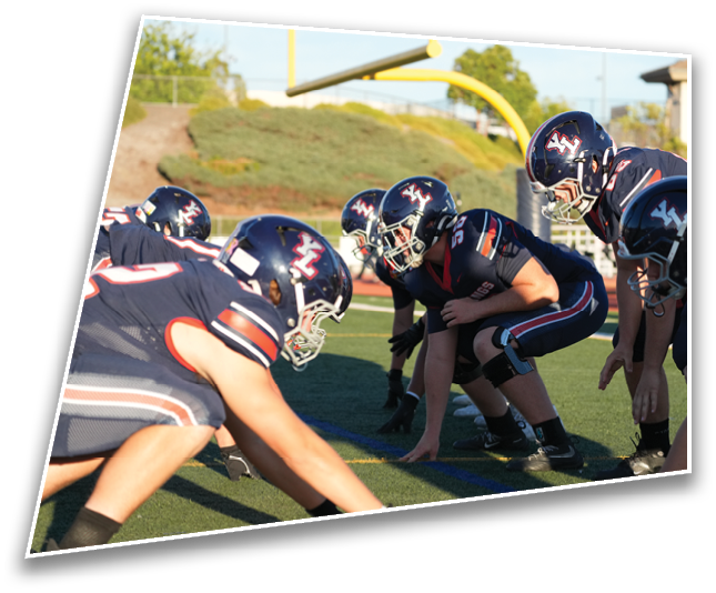 People in football uniforms and helmets are crouched in a line on a field preparing for a play under a clear sky. Description generated by AI