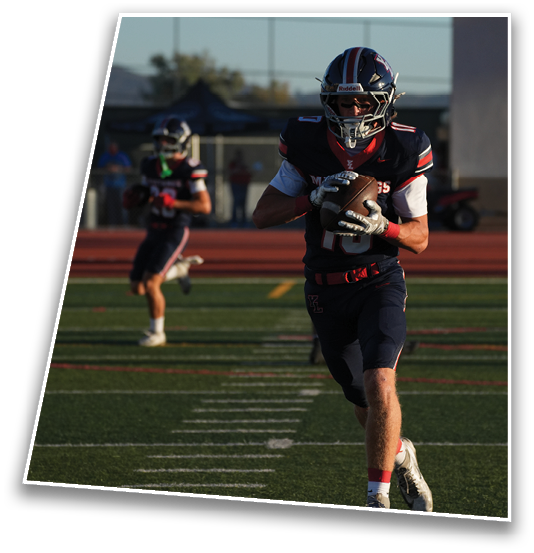 A person in a dark football uniform is running on a field while holding a football, with another person in a similar uniform running in the background. Description generated by AI