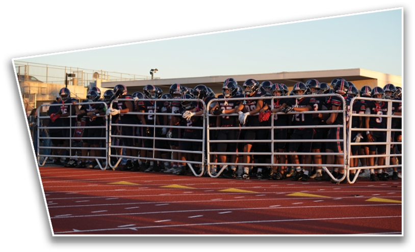 A large group of people wearing football uniforms and helmets stand closely together behind a metal barrier on a sports field. Description generated by AI