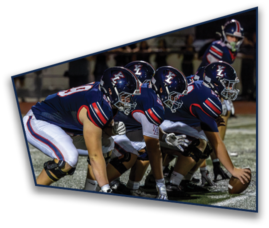 Four football players in navy blue uniforms and helmets are lined up on a field with one holding a football, preparing for a play. Description generated by AI
