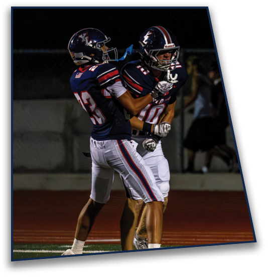 Two people wearing football uniforms are standing close together on a field at night. Description generated by AI