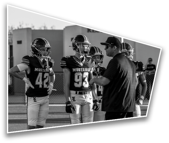 A group of people wearing football uniforms with “Mustangs" on the jerseys are standing and listening to a person in a cap and sunglasses who appears to be giving instructions near a building labeled "Restrooms." Description generated by AI