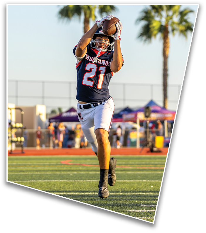A person wearing a football uniform with the number 21 is catching a football on a field with palm trees and tents in the background. Description generated by AI