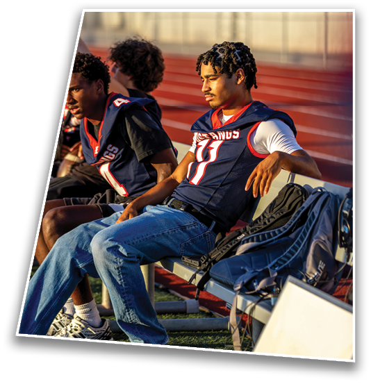 Three people wearing football jerseys sit on a bench by a track, with one person in jeans and a white undershirt looking thoughtful in the sunlight. Description generated by AI