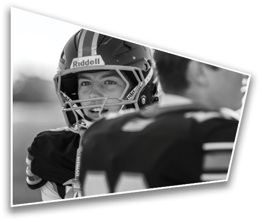 Two people wearing football helmets and uniforms are engaged in conversation on a field, with one person smiling. Description generated by AI