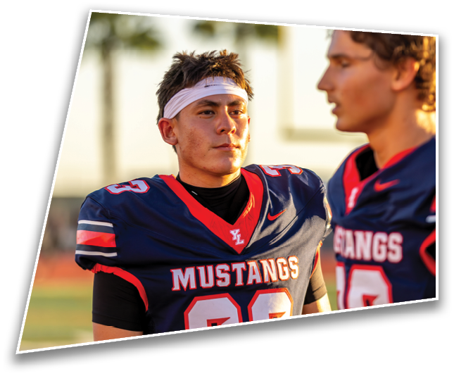 Two people wearing navy blue football uniforms with “MUSTANGS" written on the front are standing outdoors, one looking ahead with a focused expression and a white headband. Description generated by AI