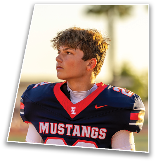 A person wearing a dark blue and red Mustangs football jersey looks to the side while standing outdoors on a field. Description generated by AI