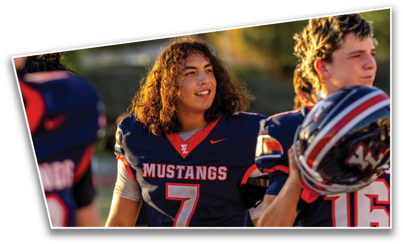 People in football uniforms with “Mustangs" on the jerseys are standing together, one holding a helmet and another smiling. Description generated by AI