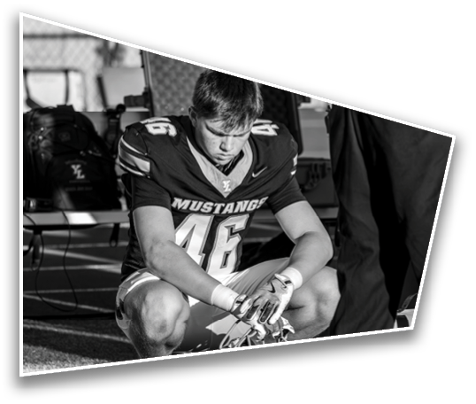A person wearing a football uniform with the number 46 and the word Mustang is crouched down with their head bowed and hands clasped, appearing to be in a moment of reflection or prayer on the sidelines. Description generated by AI