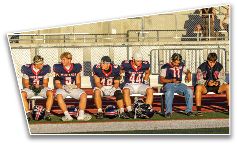 Six people wearing football jerseys and casual clothing sit on a bench at a sports field with helmets on the ground in front of them. Description generated by AI