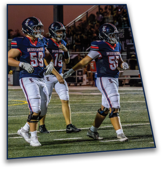 Three people in football uniforms with helmets and knee braces walk together on a field during a game at dusk. Description generated by AI