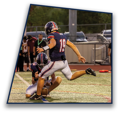 A person in a football uniform is kicking a ball held by another person kneeling on a football field during a game. Description generated by AI