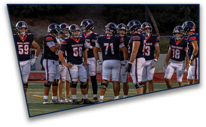 A group of people wearing football uniforms with helmets stand together on a field during what appears to be a team huddle or discussion. Description generated by AI
