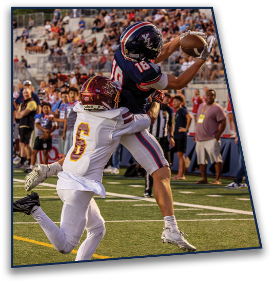 Two people in football uniforms are competing to catch a football on a field with spectators in the background. Description generated by AI