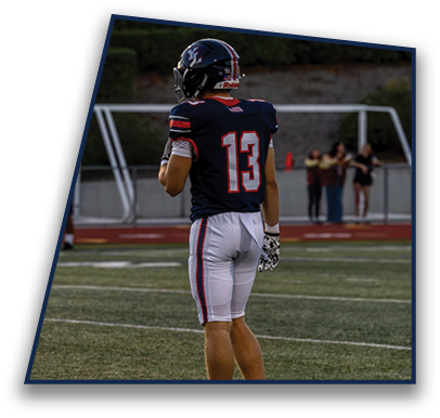 A person wearing a dark football uniform with the number 13 on the back is standing on a football field holding a football near a goalpost with blurred people in the background. Description generated by AI