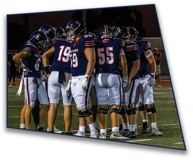A group of football players in navy blue jerseys and white pants are huddled together on a field during a nighttime game. Description generated by AI