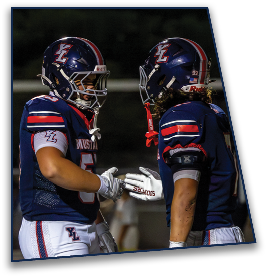 Two people wearing blue football uniforms and helmets are facing each other and appear to be about to high-five or shake hands on a field at night. Description generated by AI