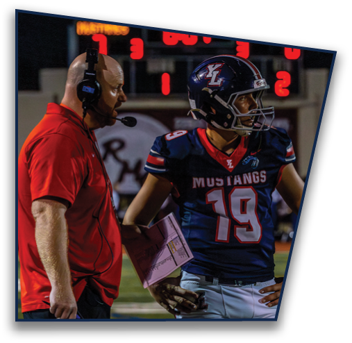 Two people on a sports field are engaged in conversation, one wearing a red shirt and holding a clipboard, and the other in a dark jersey with white pants and a wristband. Description generated by AI