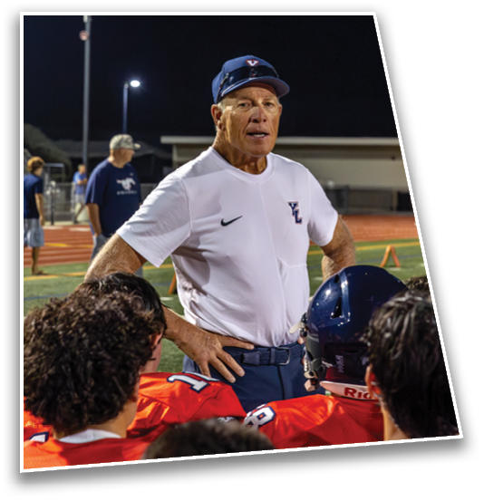 A senior person wearing a white sports shirt and cap is standing and talking to a group of people in red sports uniforms on a football field at night. Description generated by AI