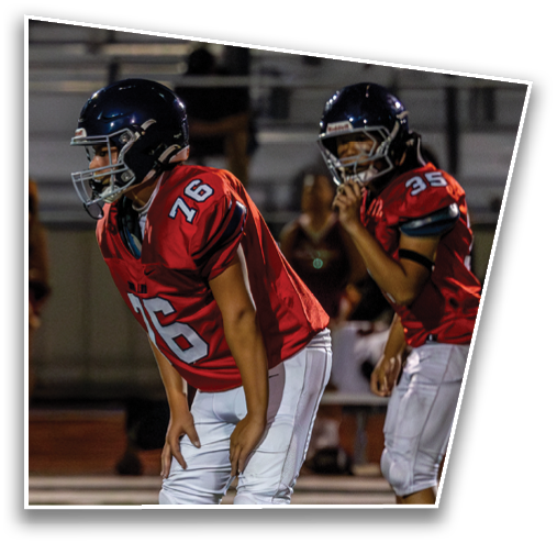 Two people wearing red football jerseys and helmets are positioned on a field, appearing ready for action during a game. Description generated by AI