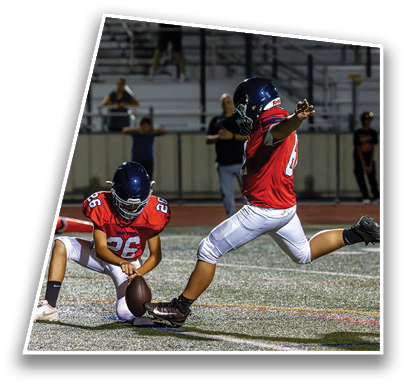 Two people in red football uniforms and helmets are on a field with one holding the ball while the other prepares to kick it. Description generated by AI
