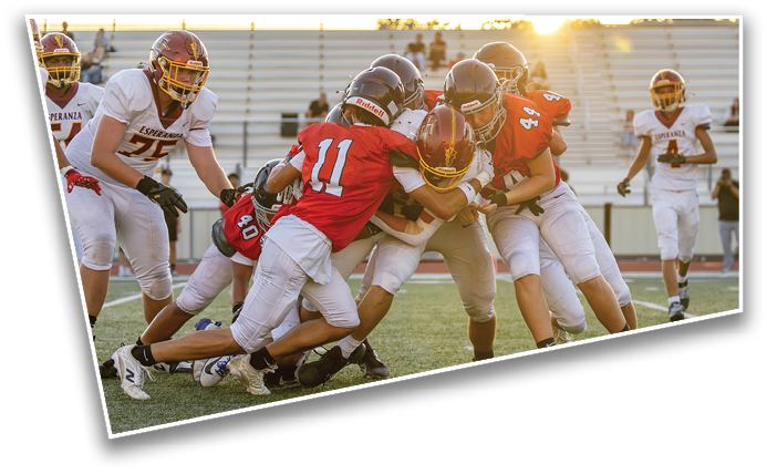 People wearing football uniforms are engaged in a tackle on a field with empty bleachers and sunlight in the background. Description generated by AI