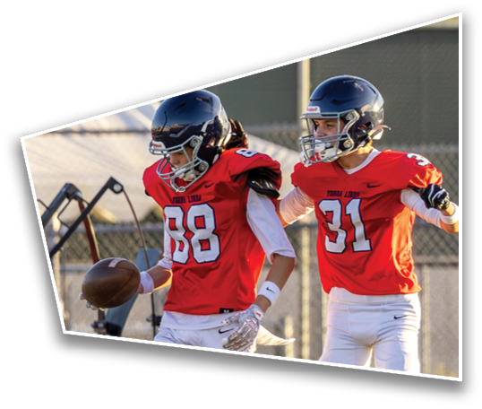 Two people wearing helmets and red football jerseys are running outdoors near a fence with trees in the background. Description generated by AI