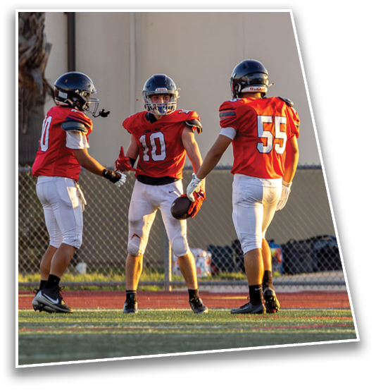 Three people wearing red football jerseys and helmets stand on a field, appearing to celebrate or communicate during a game. Description generated by AI