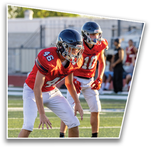Two adults in red football jerseys and helmets are crouched in a ready position on a football field during daylight. Description generated by AI
