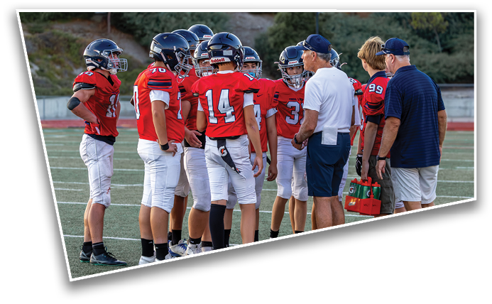 A group of people in football uniforms and helmets are gathered on a field with two adults in casual clothing talking to them, one holding a water bottle carrier. Description generated by AI