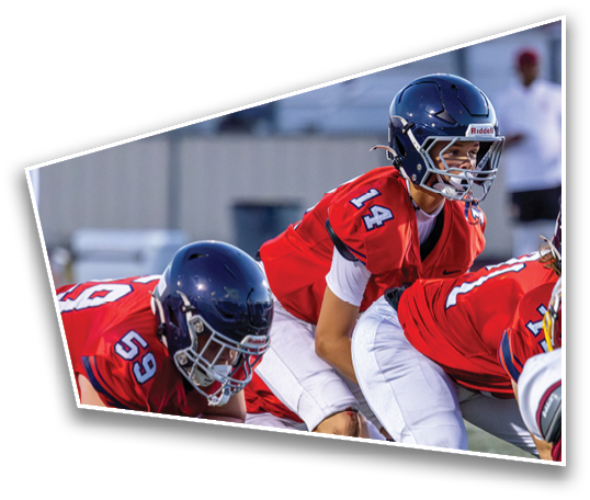 Three people wearing red football uniforms and helmets are positioned on a football field, appearing ready for a play. Description generated by AI
