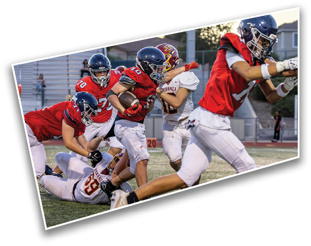 People in football uniforms are actively engaged in a game on a field with one person running with the ball while others attempt to tackle or block. Description generated by AI