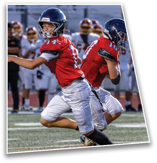 Two people in red football jerseys and helmets are actively playing on a football field with others in the background. Description generated by AI