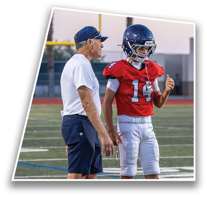 A person in a white shirt and navy shorts is talking to a person in a red football jersey and white pants wearing a blue helmet on a sports field. Description generated by AI