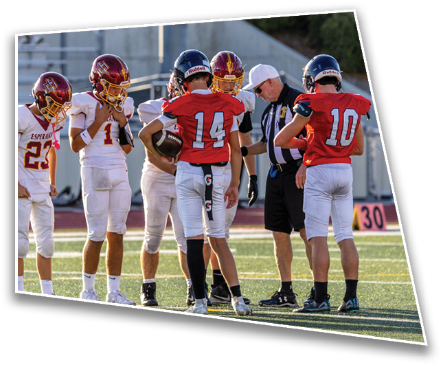 Several people in football uniforms and helmets stand on a field while a referee in a white cap checks something with them. Description generated by AI