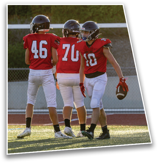 Four people wearing sports uniforms stand on a grassy field near a running track, with one holding a football. Description generated by AI