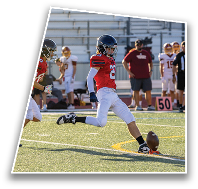 An adult in a red jersey and white pants is kicking a football on a turf field during a game with other players and a yard marker visible in the background. Description generated by AI
