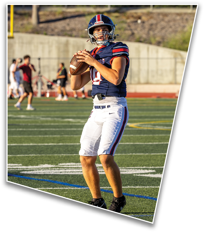 A person wearing a football helmet and uniform is holding a football in a ready-to-throw position outdoors with a blurred background of trees and a fence. Description generated by AI